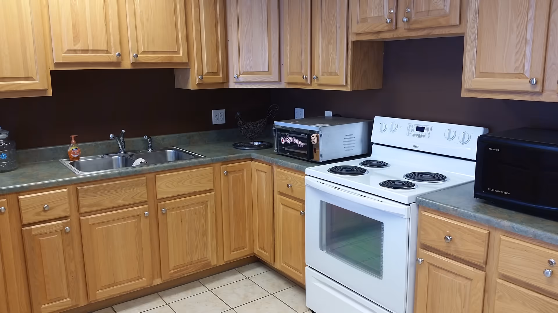A kitchen with wooden cabinets, a double sink, a white electric stove with four burners, a toaster oven, and a black microwave on the countertops. The walls are painted dark brown and the floor is tiled.