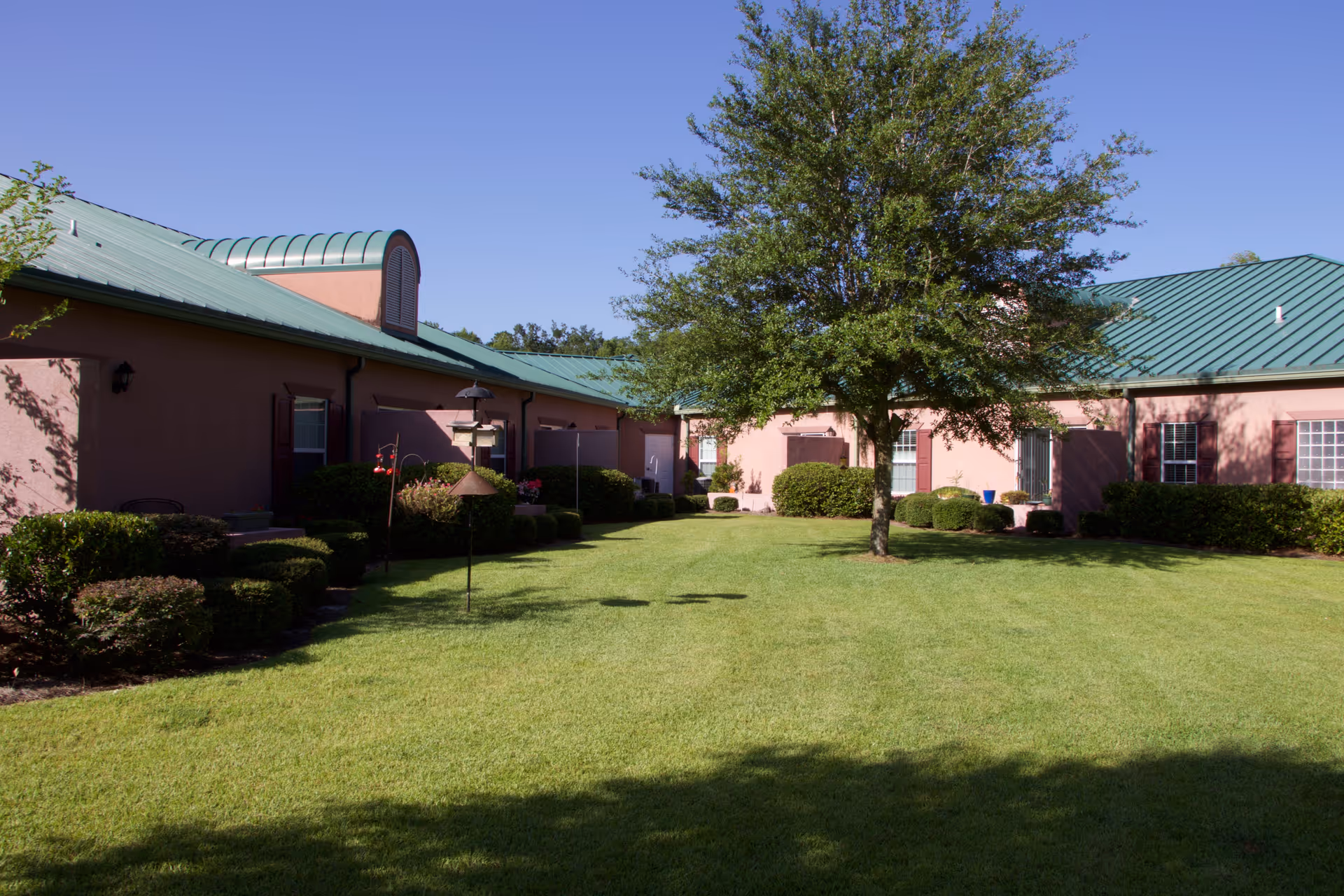 A well-maintained outdoor courtyard area with green grass, trimmed bushes, and a large tree in the center. Surrounding the courtyard are single-story buildings with green metal roofs and pinkish walls, featuring windows with shutters. The sky is clear and blue.