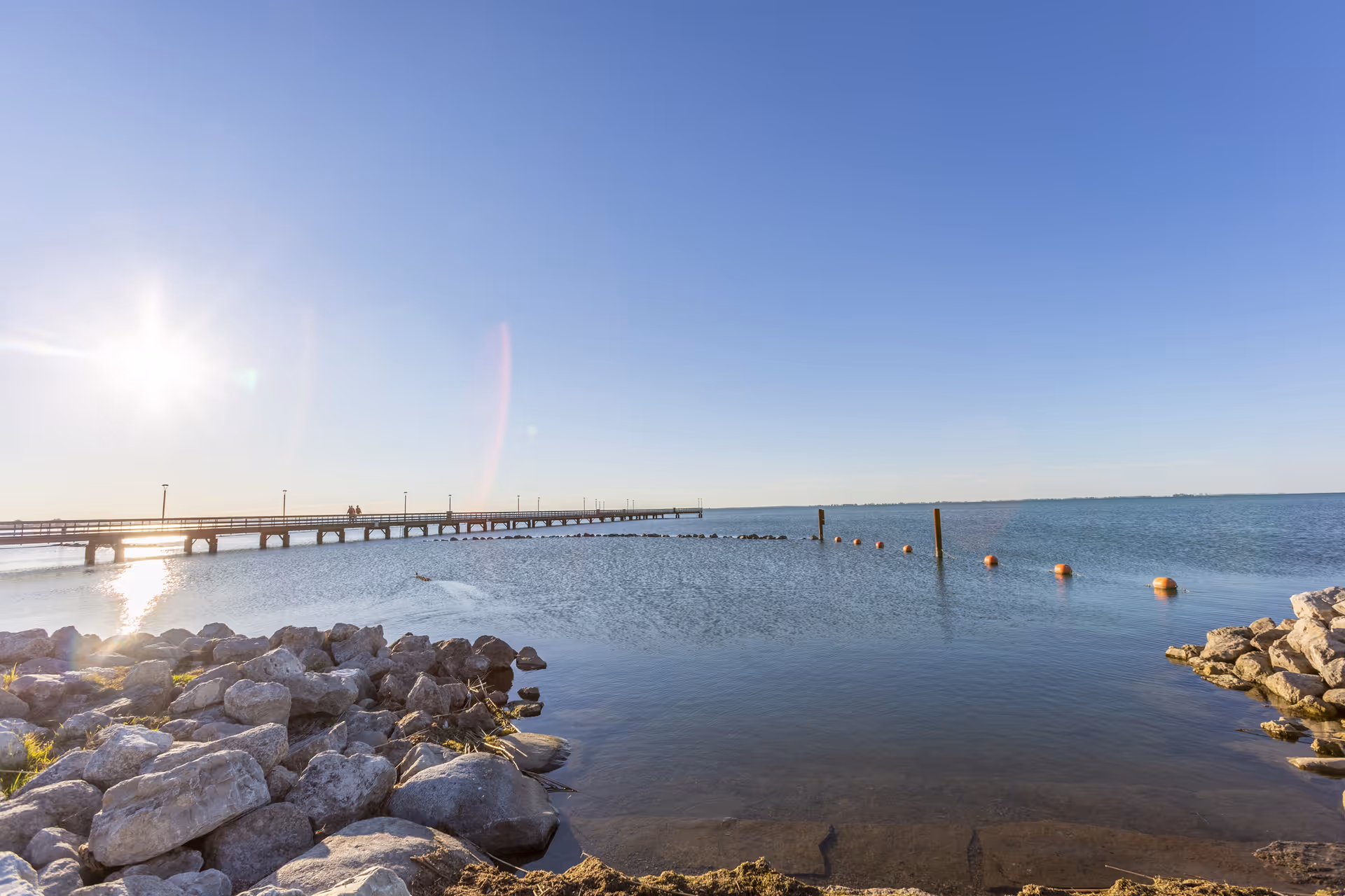 Sunlit waterfront with rocky shoreline, a long pier extending into calm water and buoys under a clear blue sky.