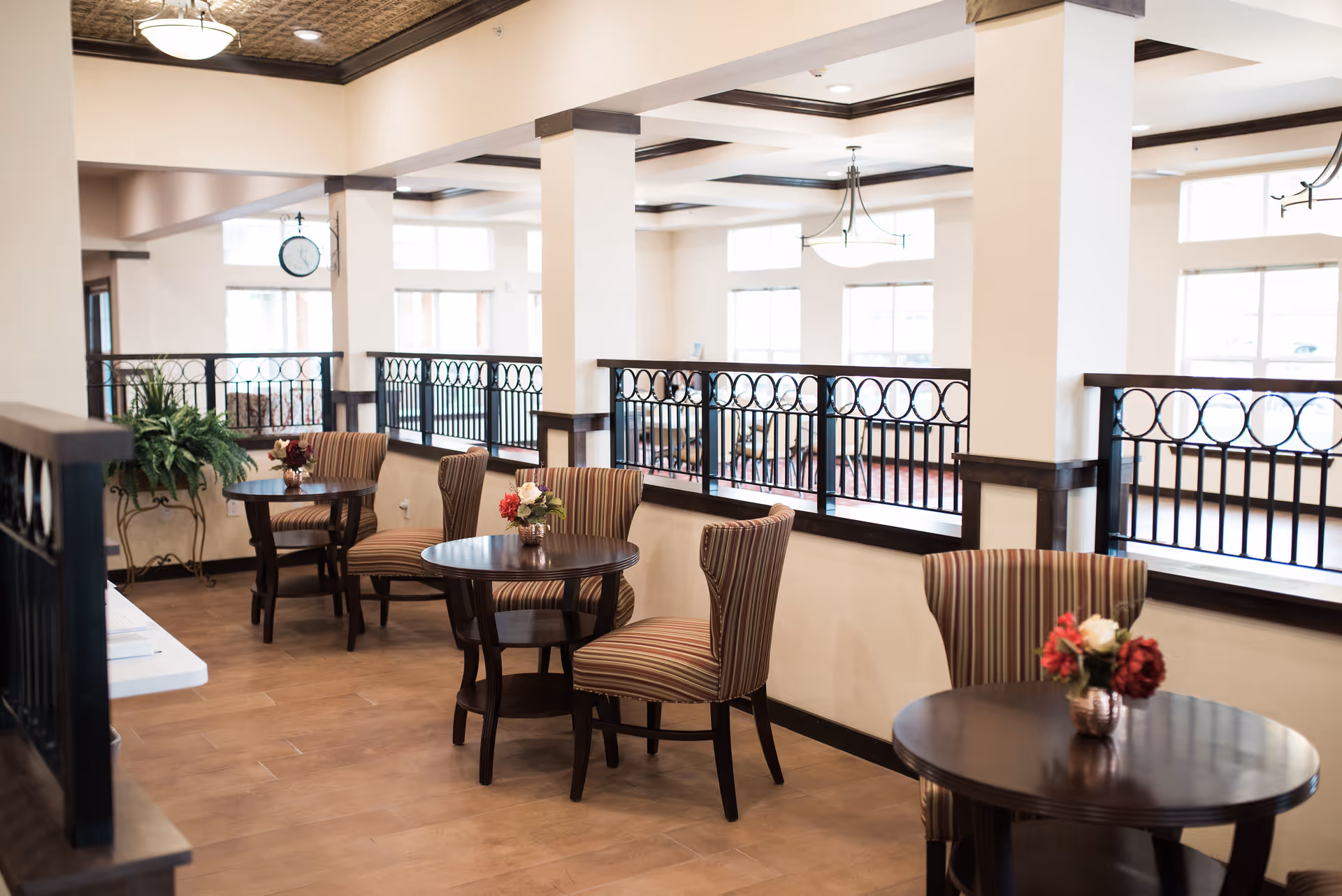 Bright communal seating area with round wooden tables, striped chairs, floral centerpieces and decorative railings in a senior living facility.
