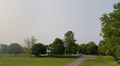 Wide view of a grassy campus with a white building and tree-lined driveway.