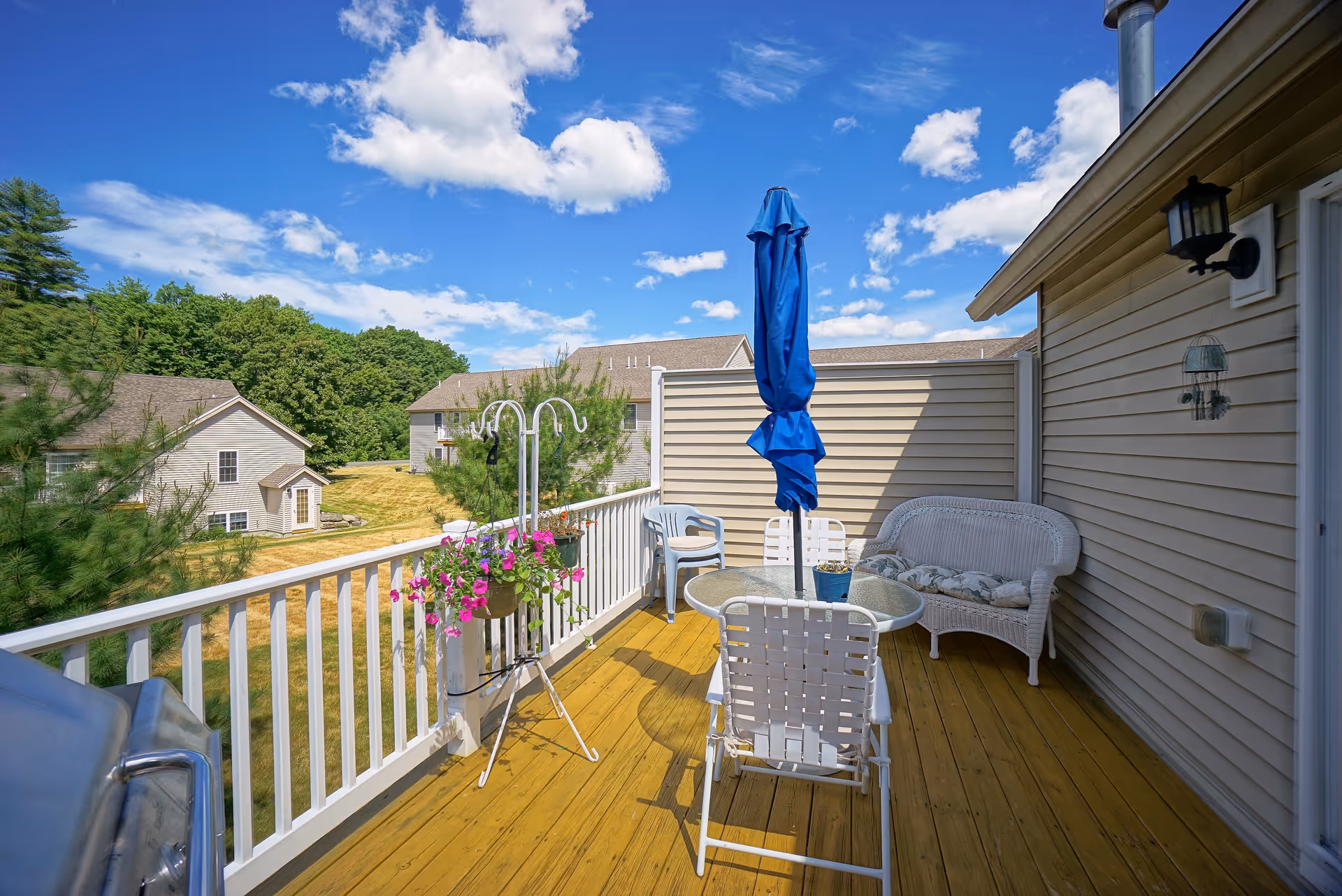 Outdoor wooden deck with white railing, a glass-top table with four white chairs, a blue closed patio umbrella, a white wicker loveseat with cushions, hanging flower pots with pink flowers, and a grill on the left side. The deck overlooks a grassy area with trees and neighboring houses under a blue sky with scattered clouds.