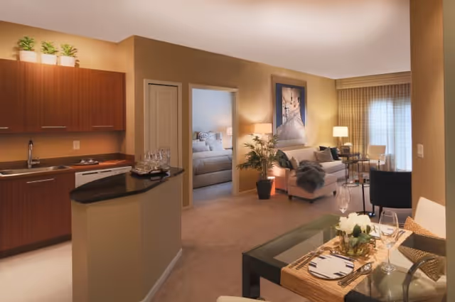 Open-plan apartment interior with a kitchenette on the left, a dining table in the foreground, and a living room with a view into a bedroom.