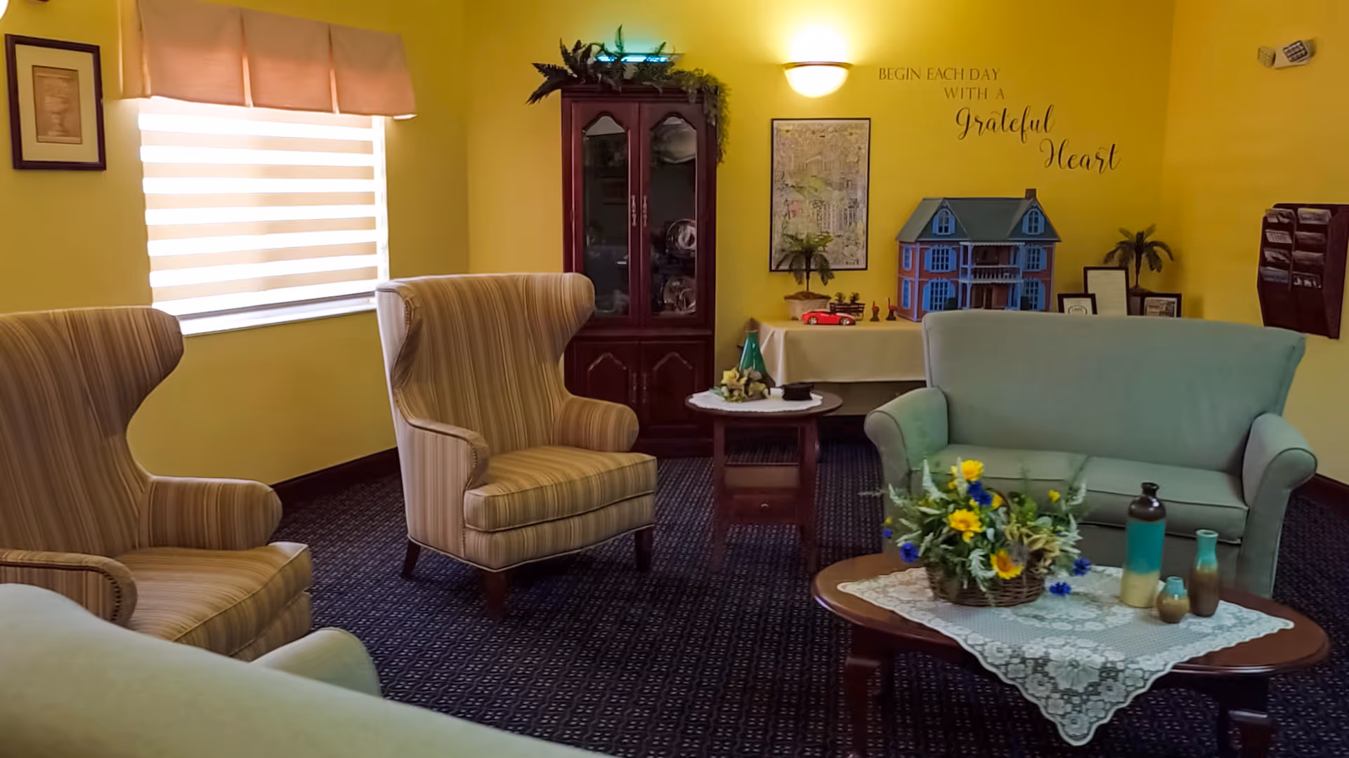 A cozy living room with yellow walls, featuring two striped armchairs, a light green loveseat, and two wooden coffee tables. One table has a basket of colorful flowers and decorative vases, while the other holds small decorative items. A wooden cabinet with glass doors stands against the wall, and a dollhouse is displayed on a table beneath a wall decal that reads 'Begin each day with a grateful heart.' A window with a beige valance and blinds lets in natural light.