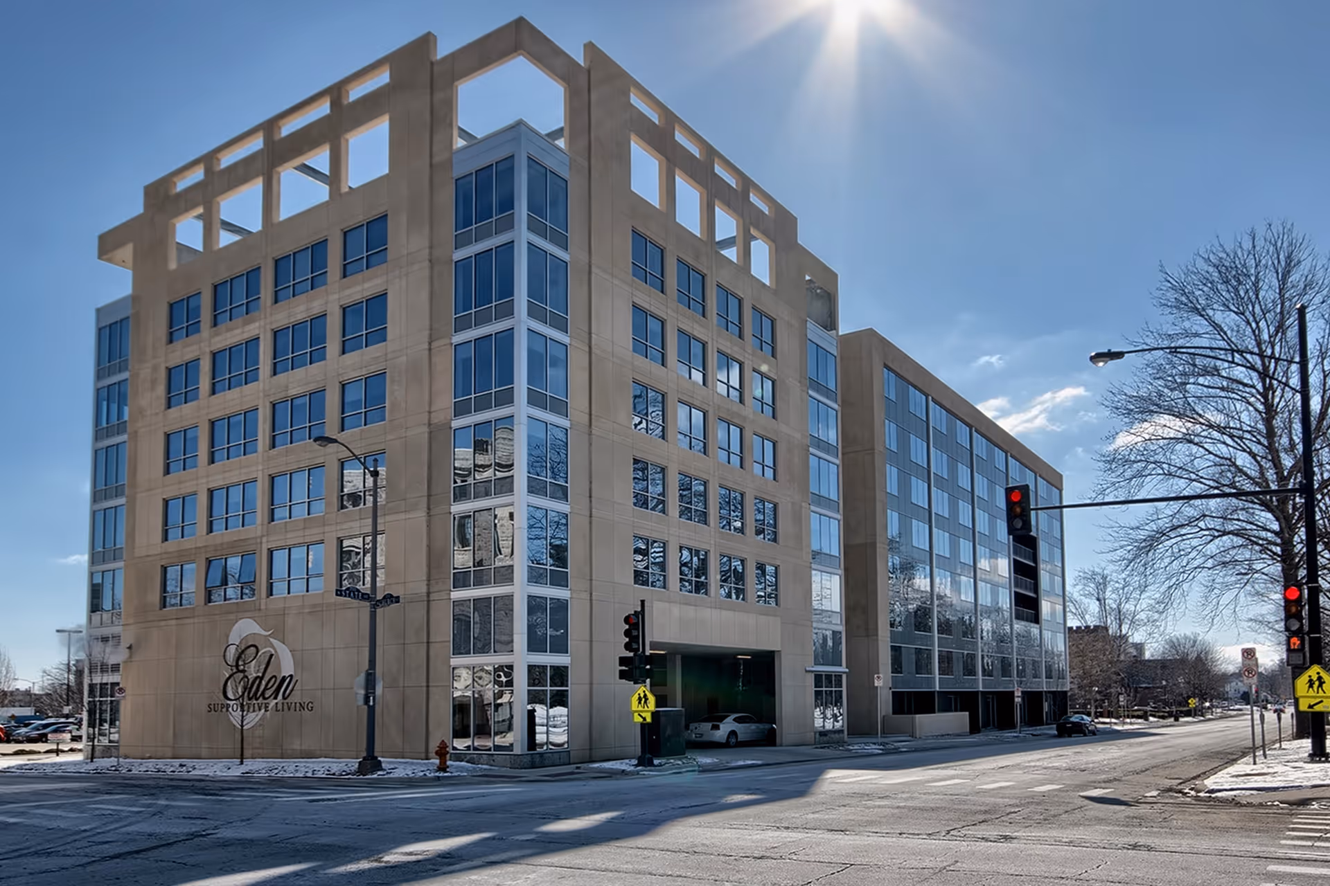 A modern multi-story concrete-and-glass building on a street corner with 'Eden Supportive Living' signage, traffic lights, and bare trees under a clear blue sky.