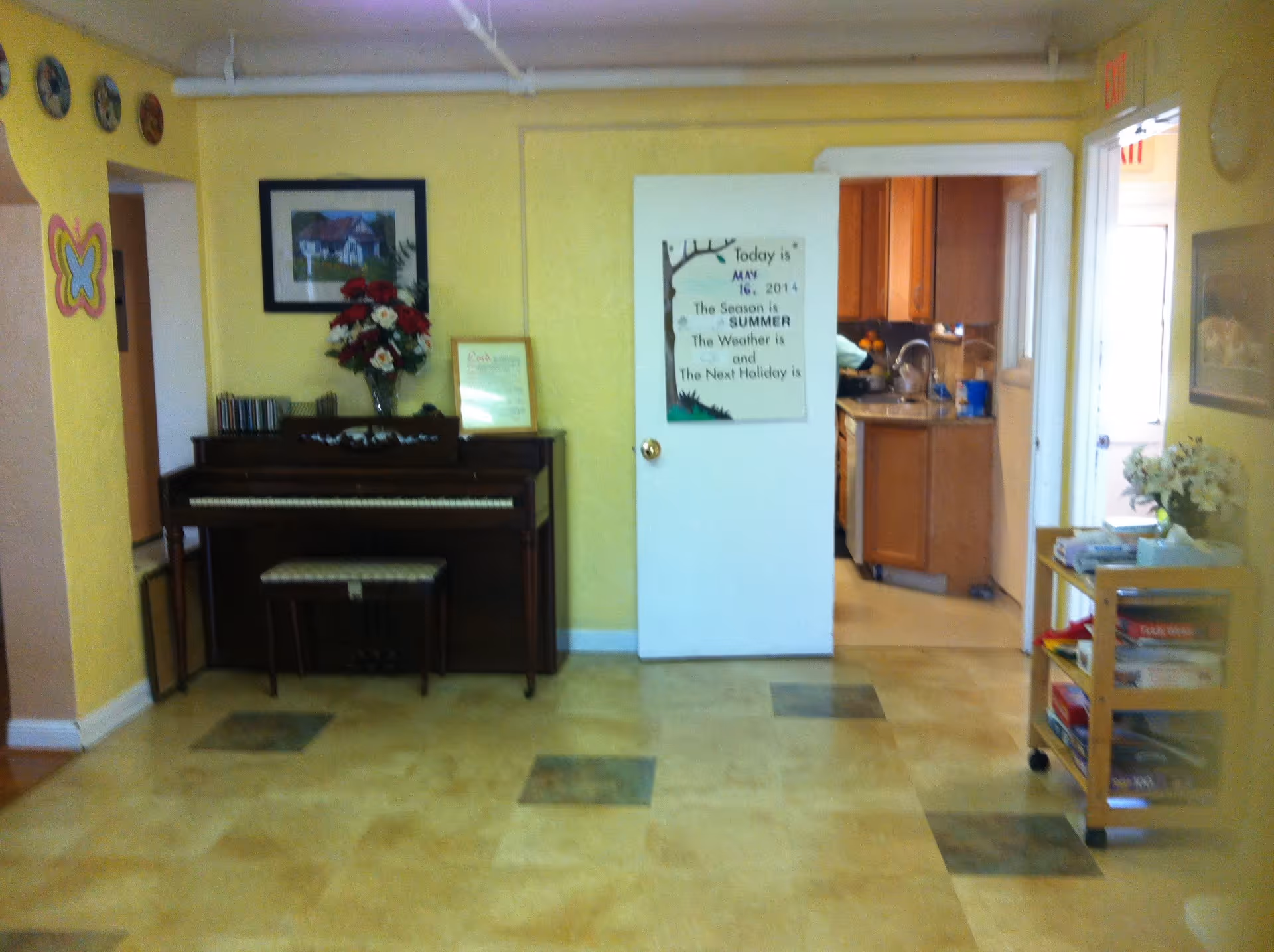 Interior room with yellow walls featuring a piano with a floral arrangement on top, a framed picture above it, and a white door leading to a kitchen with wooden cabinets. A small cart with books and flowers is on the right side of the room.
