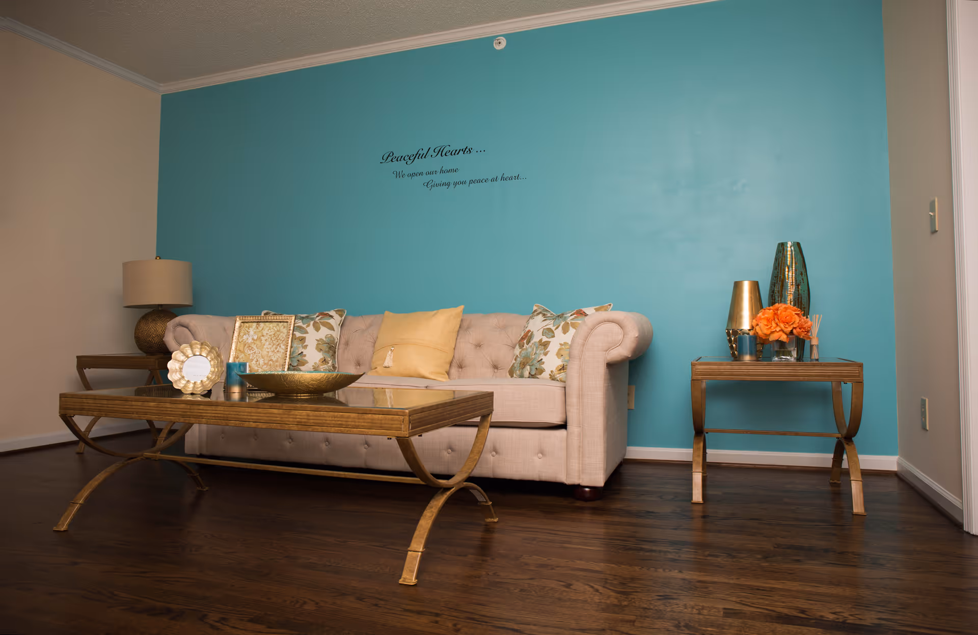 A living room with a beige tufted sofa, gold-accented coffee and side tables, decorative pillows and a teal accent wall reading 'Peaceful Hearts'.