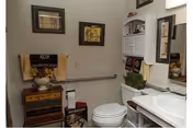A small bathroom featuring a white toilet, a white sink with a mirror above it, a small wooden cabinet with multiple drawers, decorative framed pictures on the wall, and a towel rack with a towel. The bathroom has a clean and cozy appearance with some decorative items placed on the cabinet and shelves.