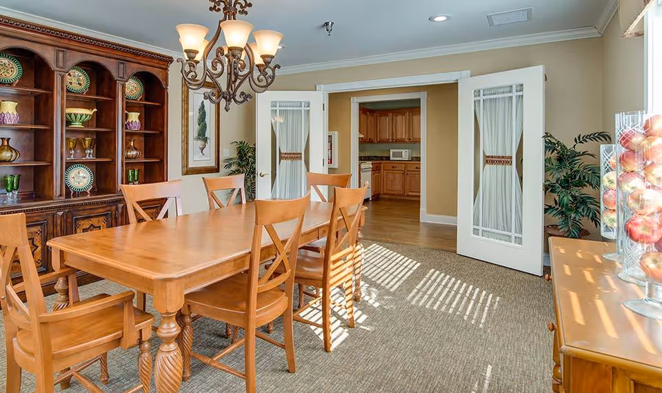 A dining room with a wooden dining table and six matching chairs. A chandelier hangs above the table. To the left, there is a wooden cabinet with decorative plates and glassware. In the background, double doors with glass panels lead to a kitchen area with wooden cabinets and a microwave. On the right, there is a sideboard with glass containers filled with apples and a potted plant next to it. Sunlight streams through a window, casting shadows on the carpeted floor.