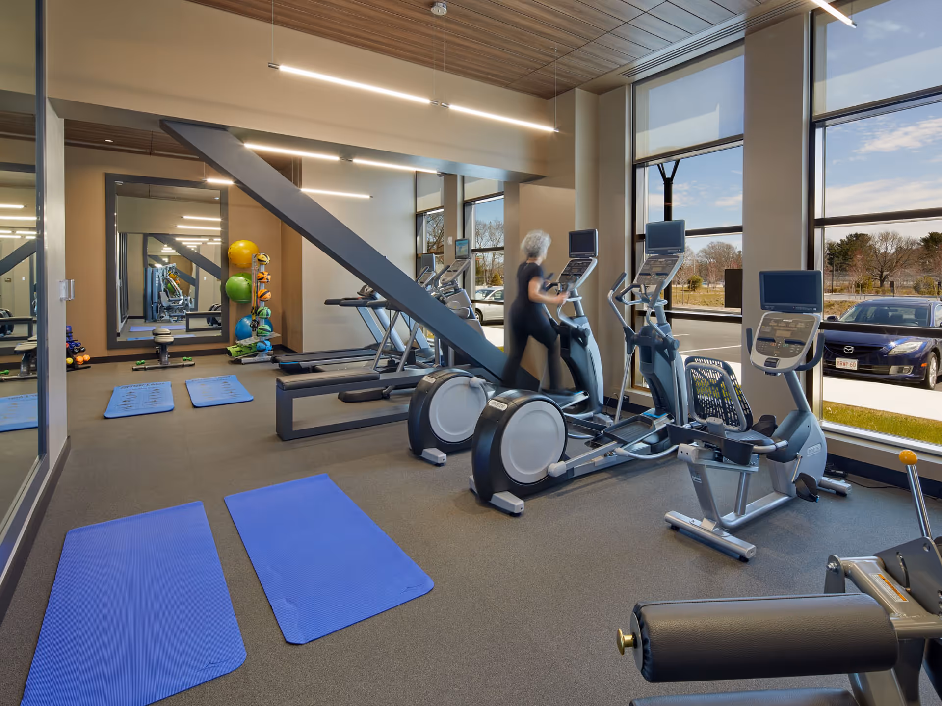 A modern fitness room with large windows showing a parking lot outside. The room contains exercise equipment including treadmills, elliptical machines, a recumbent bike, and blue exercise mats on the floor. A person with gray hair is using one of the elliptical machines. There are colorful exercise balls and weights near a large mirror on the wall.