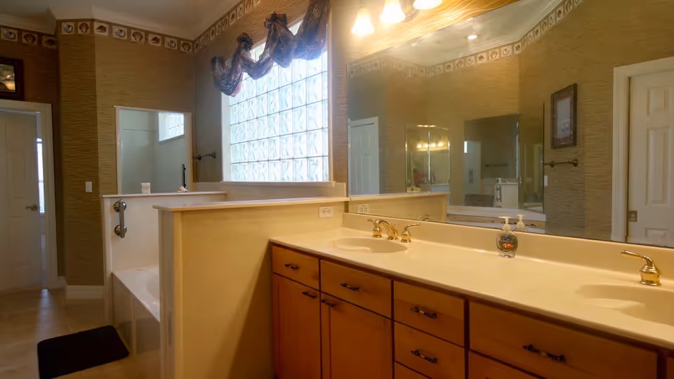 A spacious bathroom featuring a double sink vanity with wooden cabinets and gold faucets. There is a large mirror above the sinks and a frosted glass block window with a decorative valance. The bathtub is partially visible behind a half wall, and the walls have a textured wallpaper with a decorative border near the ceiling.