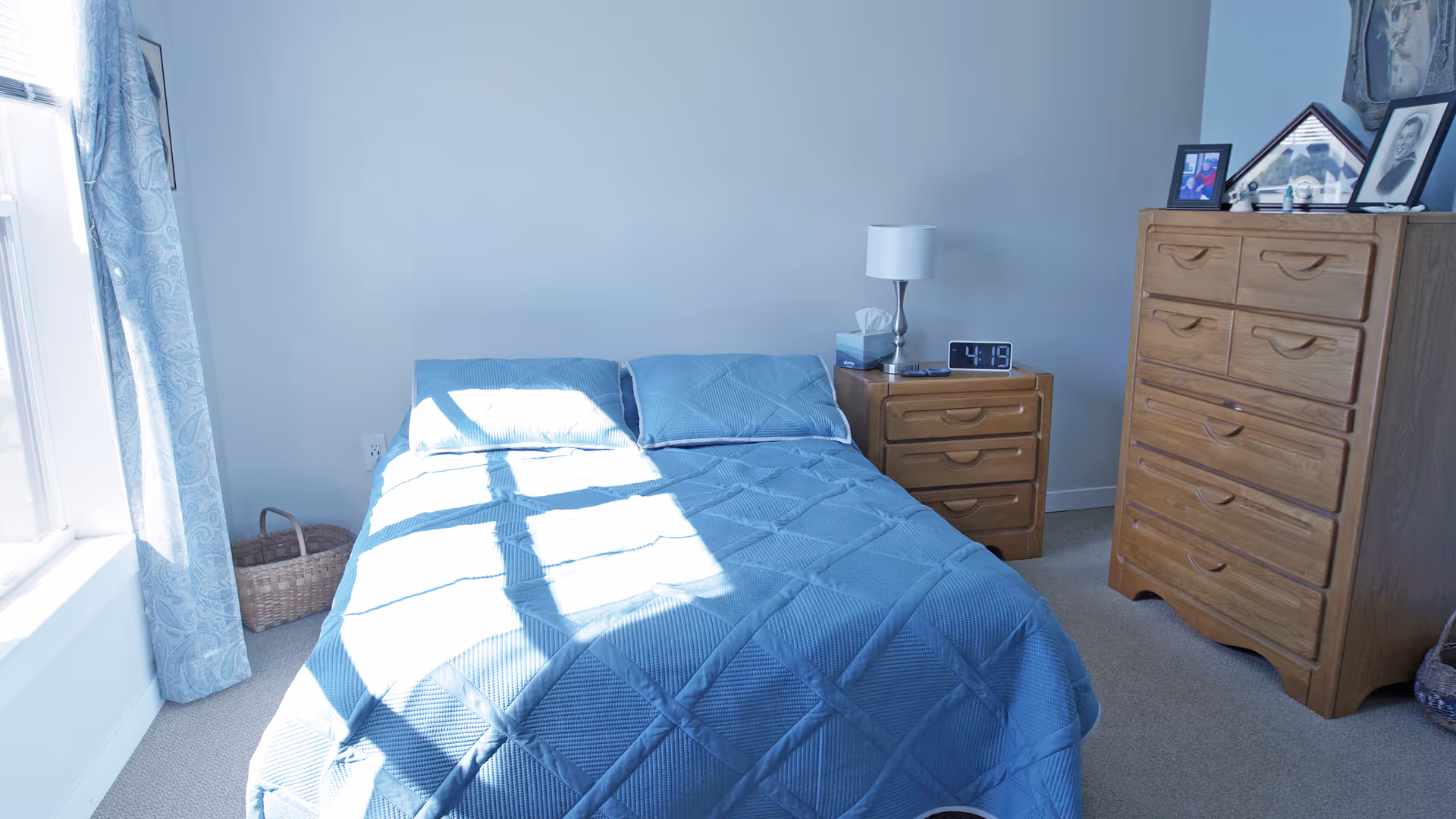 Sunlit bedroom with a blue-covered bed, a bedside table with lamp and alarm clock, and a wooden dresser.