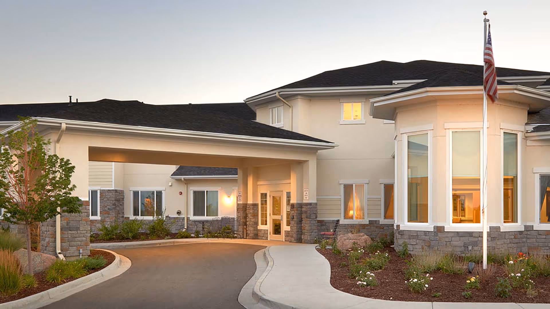 Exterior view of a senior living facility building with a covered entrance, stone and beige walls, several windows, a curved driveway, landscaped garden beds, and an American flag on a flagpole.