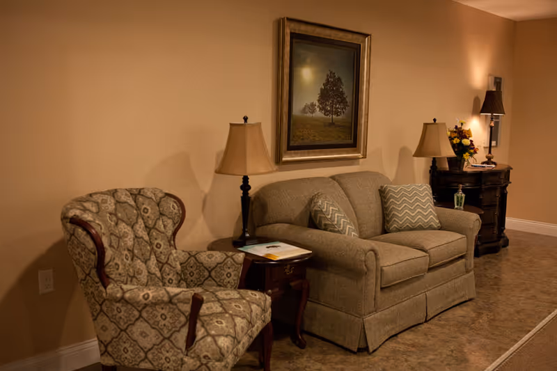A cozy living room area with a patterned armchair and a beige loveseat with two chevron-patterned pillows. Between them is a small wooden side table with a lamp and some papers. On the wall above the loveseat is a framed picture of trees in a foggy landscape. To the right is a dark wooden chest with a lamp, a flower arrangement, and a bottle of hand sanitizer. The walls are painted a warm beige color and the floor has a brownish tile.