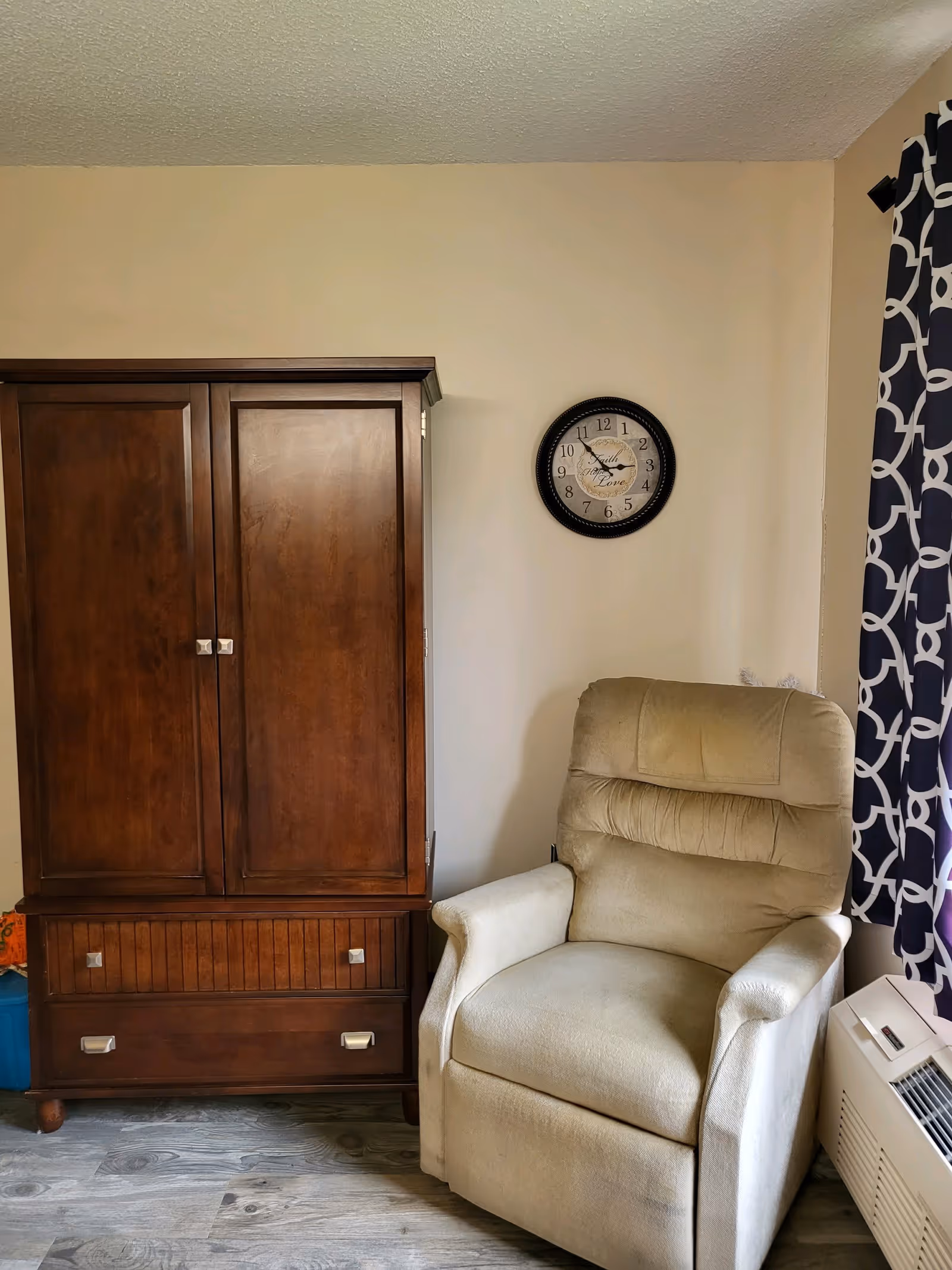 A cozy corner of a room featuring a beige upholstered recliner chair next to a dark wooden cabinet with drawers. A round wall clock hangs above the chair on a light-colored wall. To the right, there is a window with patterned curtains and an air conditioning unit below it. The floor has a wood-like finish.