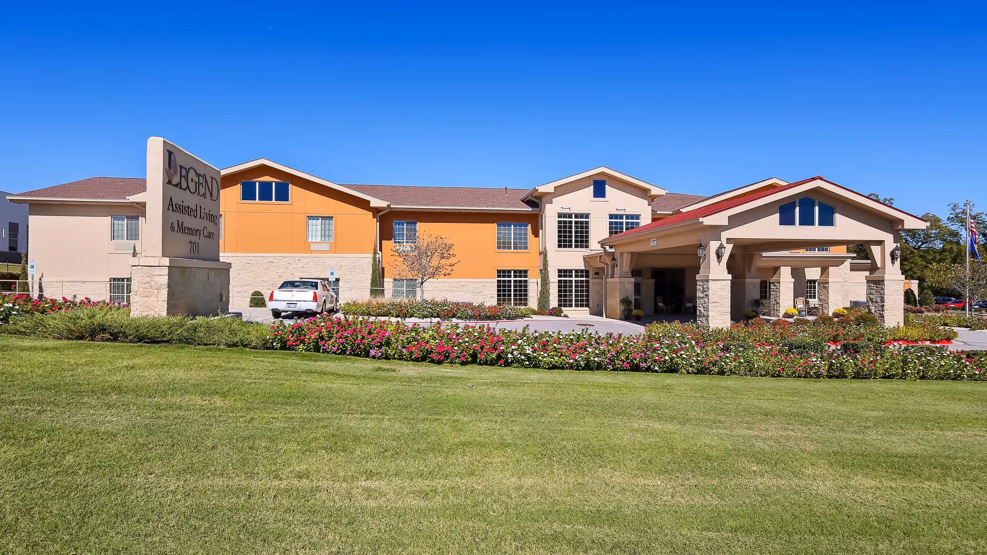 Exterior view of Legend at Tulsa Hills assisted living and memory care facility with a well-maintained lawn, flower beds, and a covered entrance under a clear blue sky.
