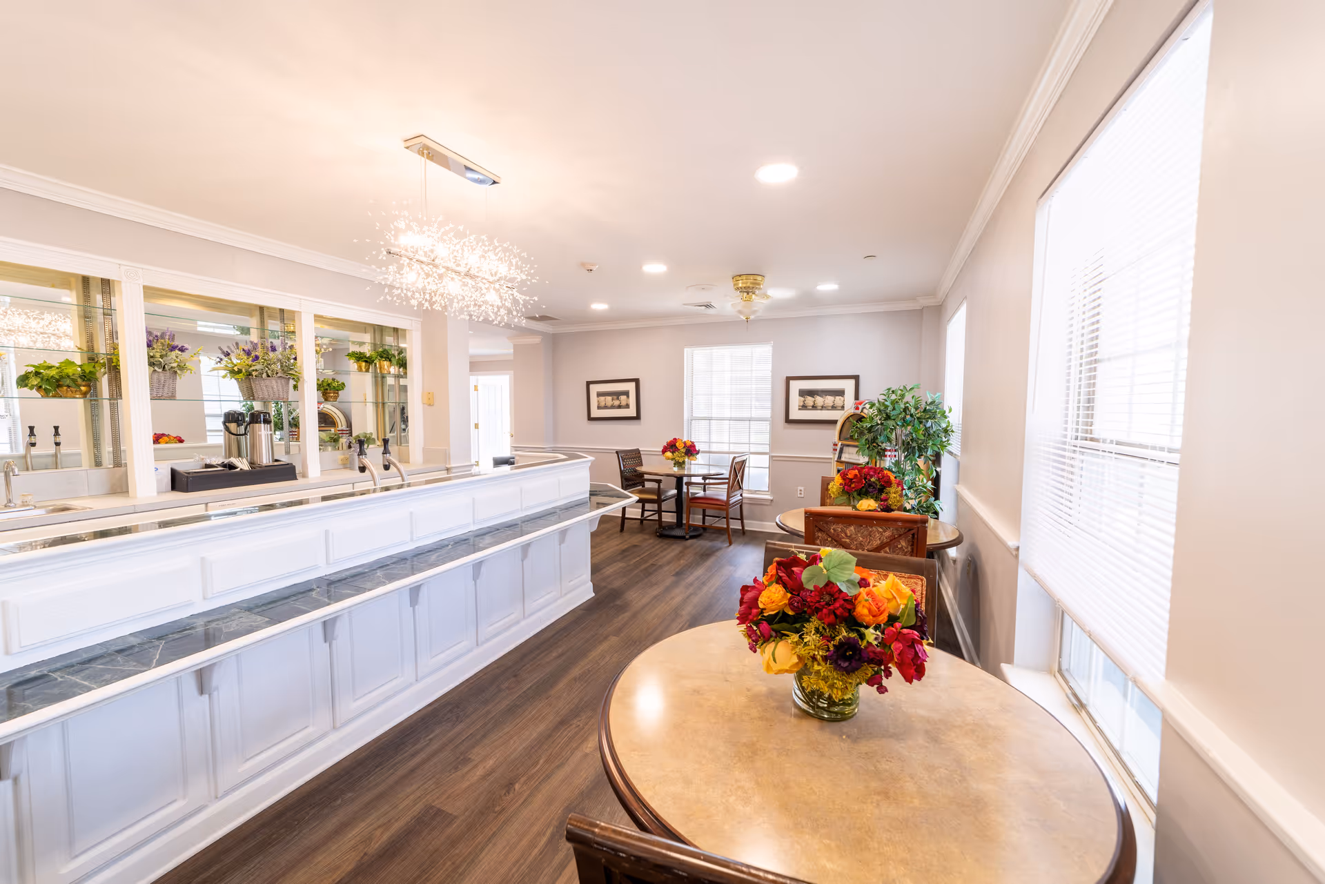 Bright dining area with round tables topped with floral centerpieces, a long white service counter, and large windows.