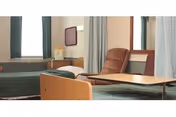 A healthcare facility room with two hospital-style beds, each with green bedding. There is a brown reclining chair and a wooden overbed table between the beds. The room has light-colored walls, a window with curtains, and a small cabinet with a mirror above it.