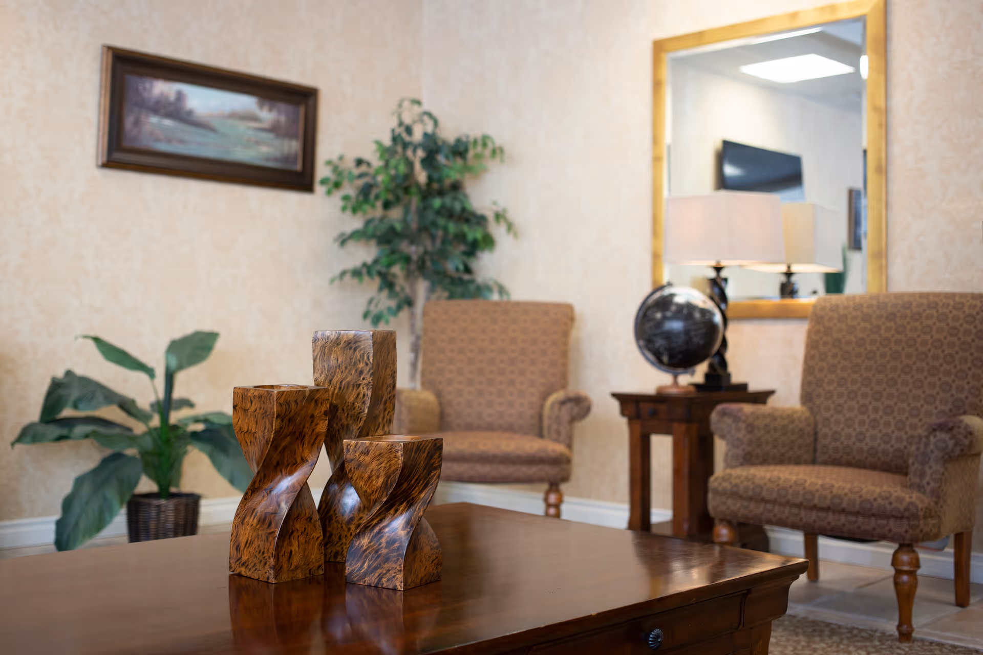 A cozy sitting area with two upholstered chairs, a wooden coffee table displaying carved wooden sculptures, a side table with a lamp and globe, a mirror, and plants.