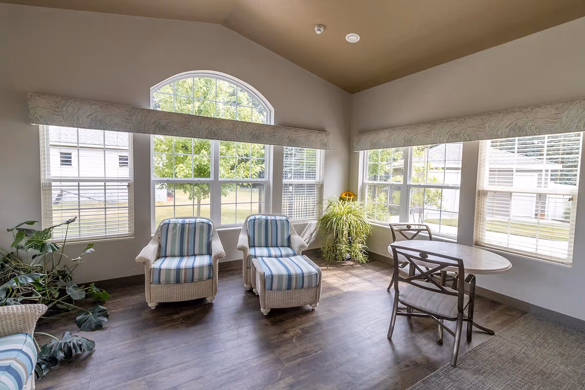 A bright sitting area with large windows showing green trees outside. The room has two striped cushioned wicker chairs with a matching ottoman, a small round table with two chairs, and several potted plants. The floor is wood with a carpeted section.