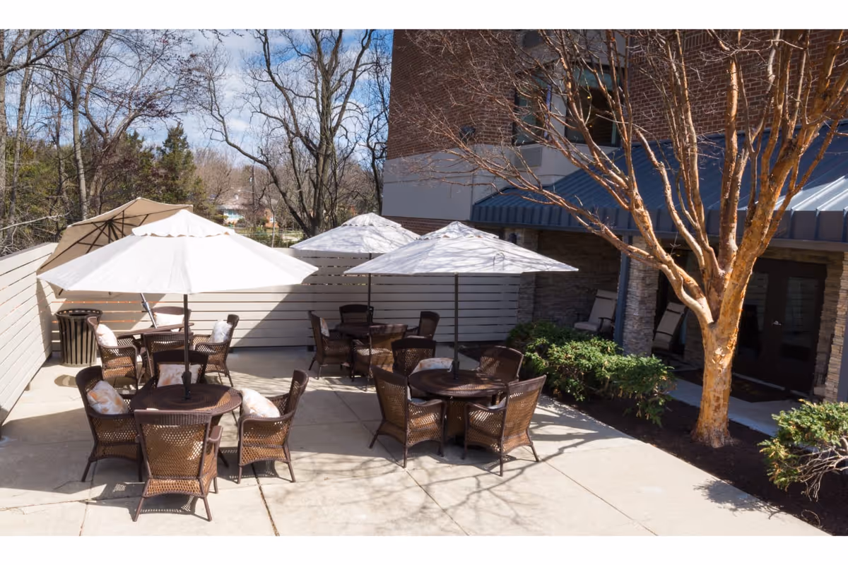 Outdoor patio area with multiple round tables and wicker chairs, each table shaded by large white umbrellas. The patio is surrounded by a wooden fence and landscaping with bushes and a tree. The building exterior with stone and brick walls is visible in the background.
