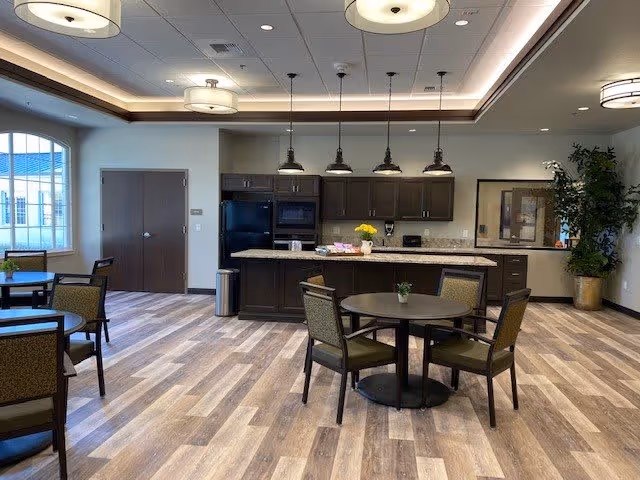 A spacious, well-lit communal kitchen and dining area with wood-look flooring, dark wood cabinets, a central island with granite countertop, and several round tables with chairs. Pendant lights hang above the island, and there is a large window on the left side letting in natural light. A potted plant is placed in the corner near a framed picture on the wall.