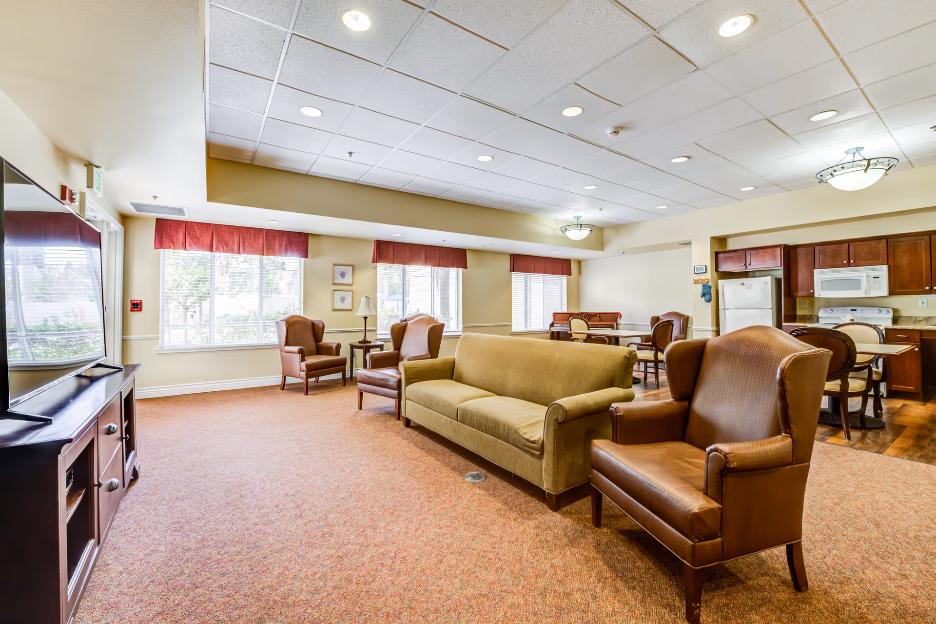 A bright and spacious senior living common area with a large TV on a wooden stand, a green sofa, and several brown armchairs. The room has large windows with red valances letting in natural light, a carpeted floor, and a ceiling with recessed lighting. In the background, there is a small kitchen area with wooden cabinets, a refrigerator, microwave, and stove, as well as a dining table with chairs.