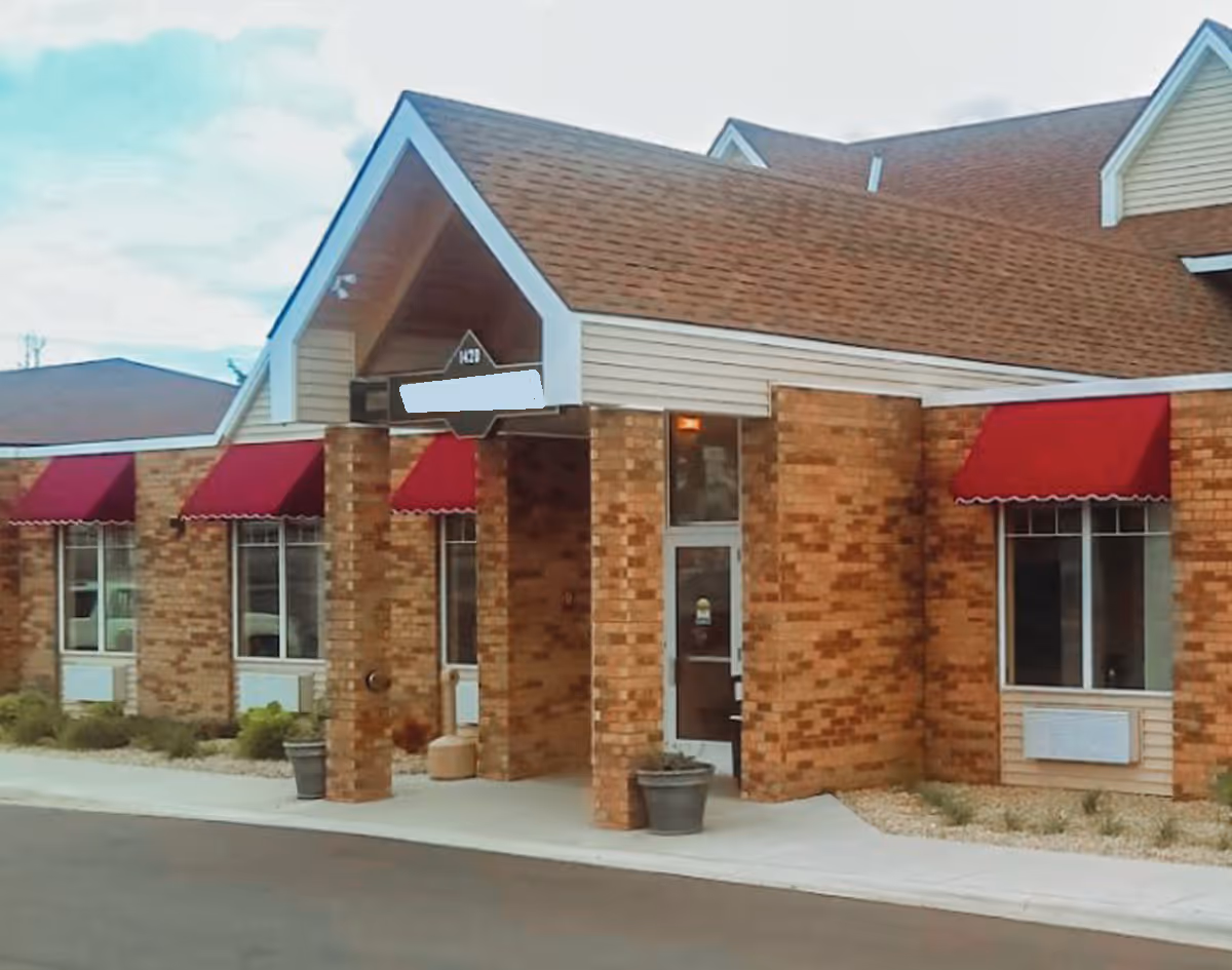 Exterior view of a brick building with a peaked roof entrance and red awnings over the windows, showing the front entrance of the facility.