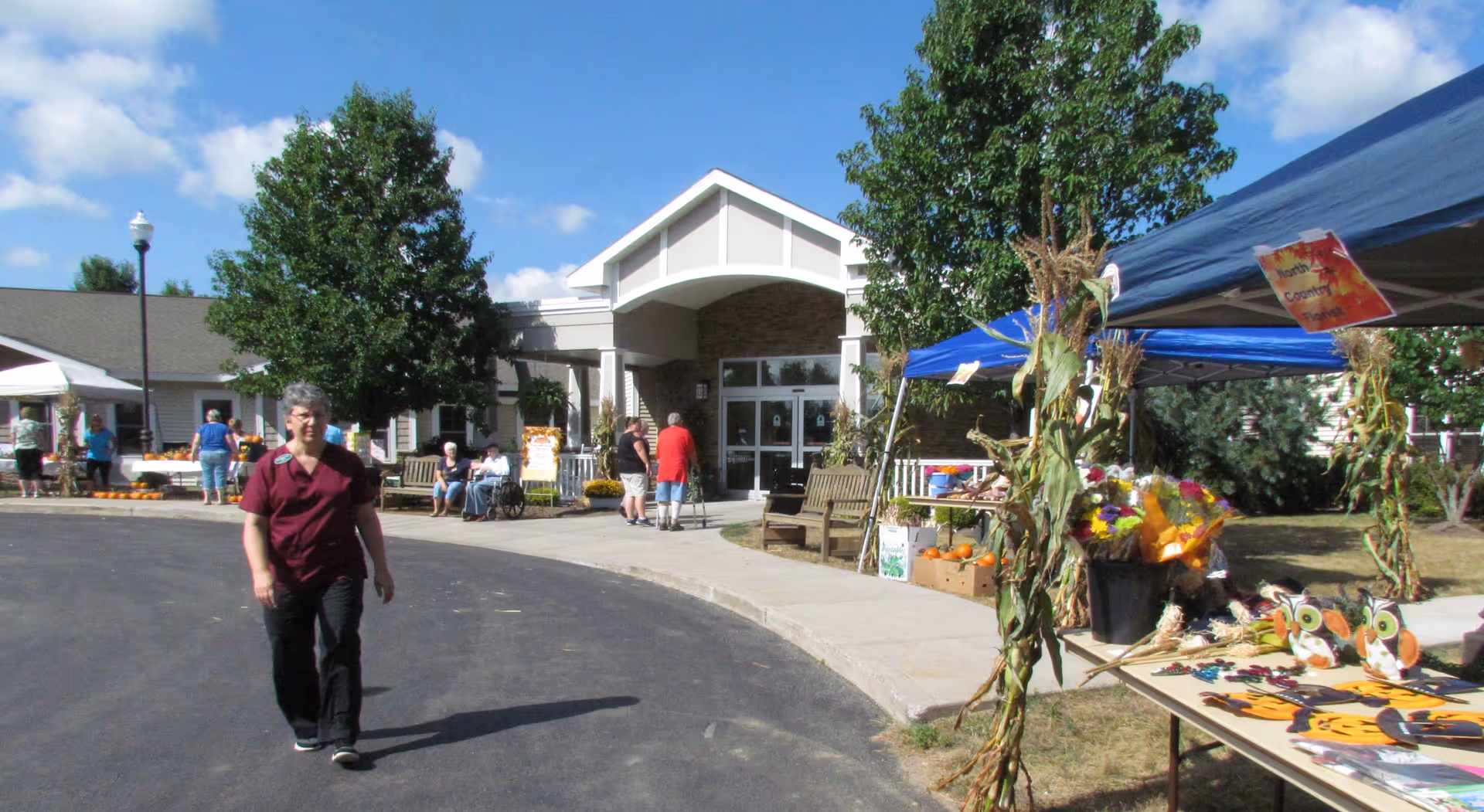 Outdoor scene at Syracuse Home at McHarrie Place showing the entrance to the building with people walking and sitting nearby. There are tents set up with tables displaying autumn-themed decorations and flowers. Trees and a clear blue sky are visible in the background.