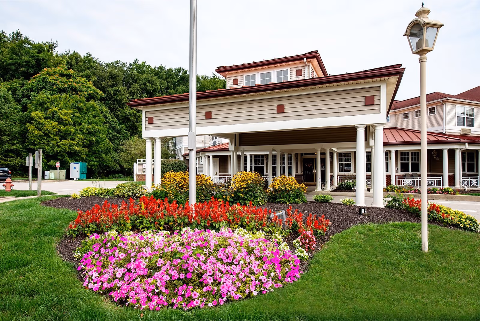 Entrance of a senior living facility with a covered driveway, surrounded by well-maintained flower beds featuring pink, red, and yellow flowers, green grass, and a lamppost on the right side. The building has beige siding with white columns and a red roof, with trees in the background.
