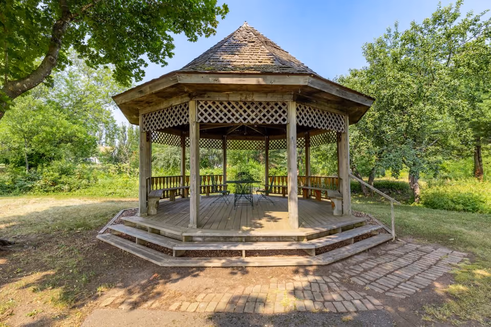 A wooden gazebo with a shingled roof situated in a green outdoor area surrounded by trees and grass. The gazebo has latticework along the top and built-in benches inside, with a small table and chairs in the center. The sky is clear and blue.