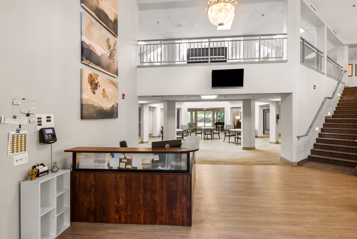 Interior view of a senior living community lobby with a wooden reception desk in the foreground, abstract paintings on the wall, a staircase on the right, and a seating area with tables and chairs in the background under a high ceiling with a chandelier.