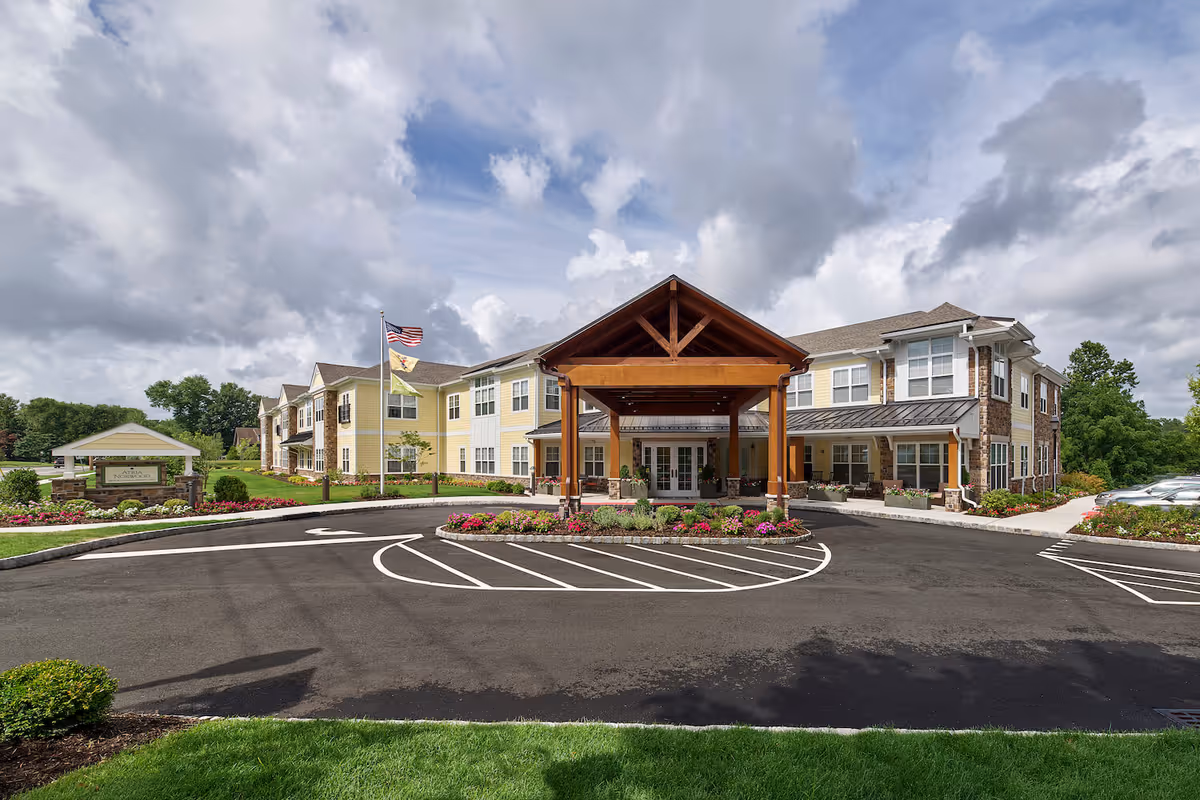 Front exterior of a two-story assisted living building with a wooden covered entrance, landscaped circular driveway, and flags.