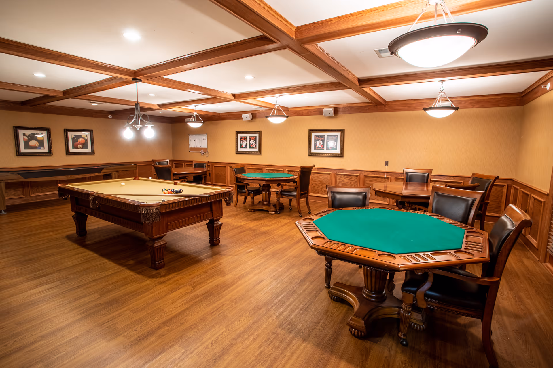 A spacious game room with wooden flooring and wood-paneled walls, featuring a pool table with balls arranged for a game, two round card tables with green felt tops, and several chairs. The ceiling has wooden beams and multiple hanging light fixtures. Framed pictures hang on the walls.