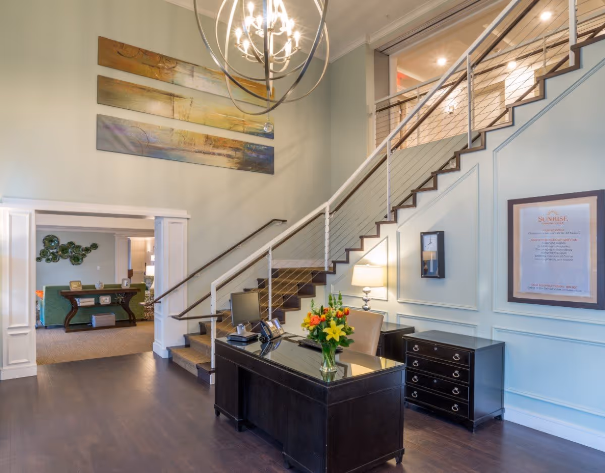 Lobby interior with a reception desk and flowers, a modern staircase with cable railing, chandelier, and seating area beyond.