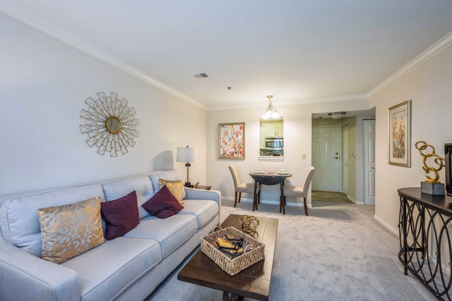 A cozy living room area with a light gray sofa adorned with gold and purple pillows, a wooden coffee table with a woven basket and decorative items, a small dining table set for two, and a view into a kitchen area. The walls are light-colored with decorative artwork and a sunburst mirror, and the floor is carpeted.