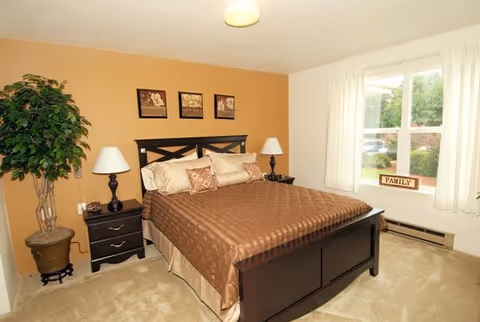 A cozy bedroom with a large dark wooden bed covered with a brown quilt and multiple pillows. There are two matching dark wooden nightstands on either side of the bed, each with a lamp. A potted plant is placed in the corner next to one nightstand. The walls are painted a warm beige color, and three framed pictures hang above the bed. A window with white curtains lets in natural light, and a small decorative sign that says 'FAMILY' is placed on the windowsill.