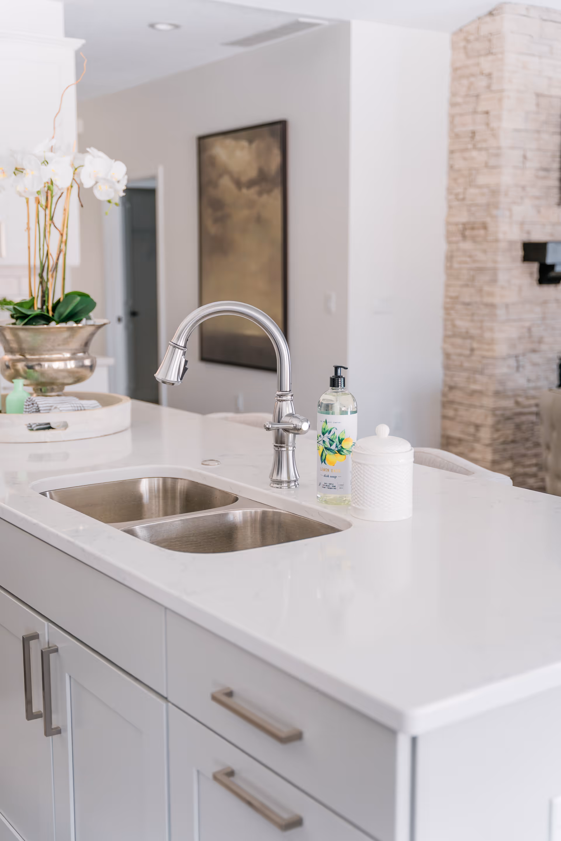 Modern kitchen island with a double stainless steel sink, a silver faucet, a bottle of hand soap, and a white decorative container on a white countertop. In the background, there is a silver vase with white orchids and a blurred view of a living area with a stone fireplace and a wall painting.