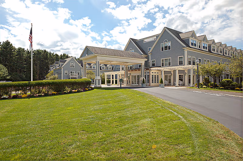 Exterior view of a large senior living facility building with gray siding and white trim, featuring multiple windows and dormers. The entrance has a covered drop-off area supported by columns. There is a well-maintained green lawn and a flagpole with an American flag on the left side. The sky is partly cloudy.