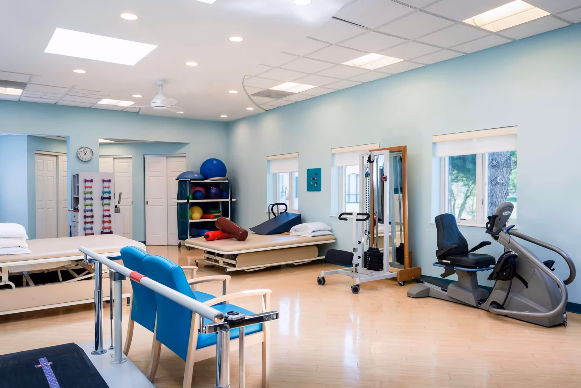 A bright physical therapy room with light blue walls and wooden flooring. The room contains two therapy beds with pillows, a rack of colorful exercise balls and foam rollers, a set of dumbbells on a rack, a parallel bar for walking exercises, a stationary recumbent exercise bike, and a multi-functional exercise machine. There are three windows letting in natural light and a clock on the wall.