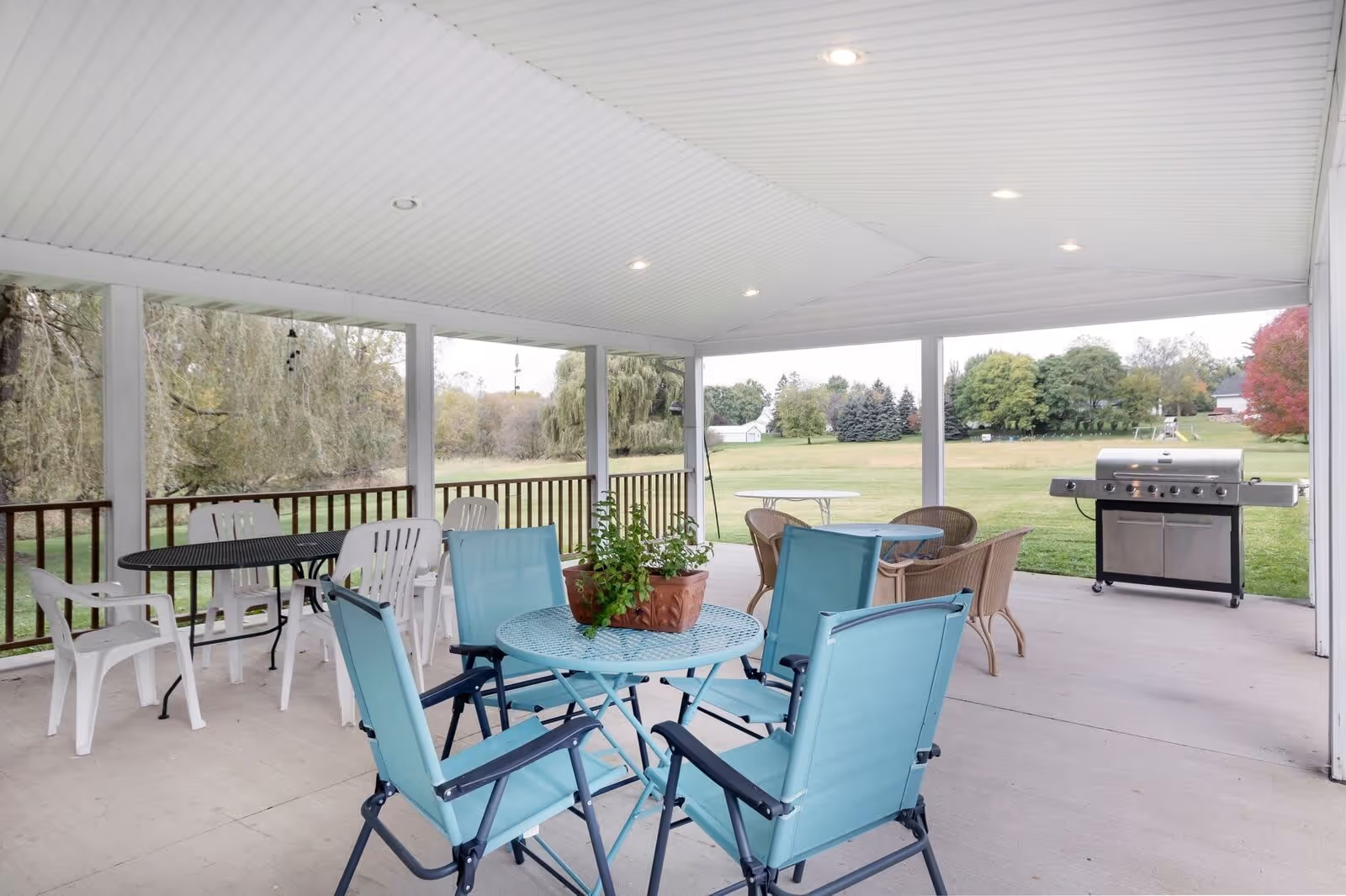 Covered outdoor patio area with multiple seating arrangements including blue metal table with four blue chairs, black metal table with white plastic chairs, and a round table with wicker chairs. There is a large stainless steel grill on the right side and a view of a grassy field with trees in the background.