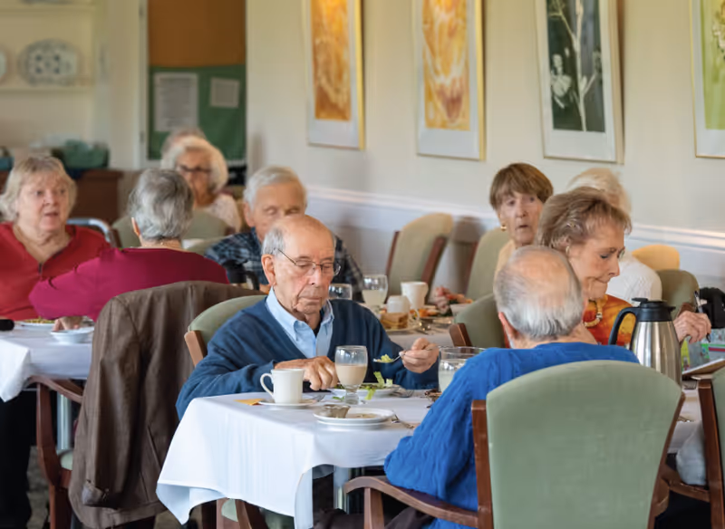 A group of elderly people sitting at tables in a dining room, eating and drinking. The room has light-colored walls with framed artwork and green cushioned chairs.