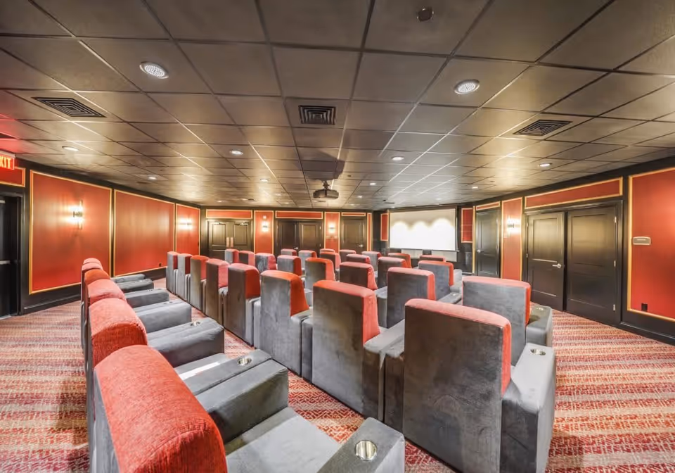 Interior view of a theater room with multiple rows of comfortable gray and red armchairs facing a large white projection screen. The room has red walls with gold trim, a patterned carpet, and a ceiling with recessed lighting and ventilation panels.