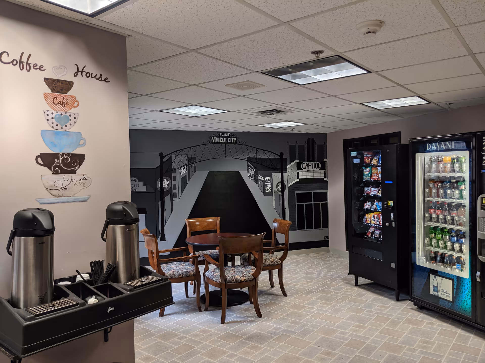 A small coffee house area inside a facility with a wall mural depicting a street scene labeled Flint Vehicle City. There is a round table with four wooden chairs with patterned cushions. On the left, there are two stainless steel coffee dispensers on a black stand with coffee supplies. On the right, there are vending machines stocked with snacks and beverages. The floor is tiled and the ceiling has fluorescent lights.