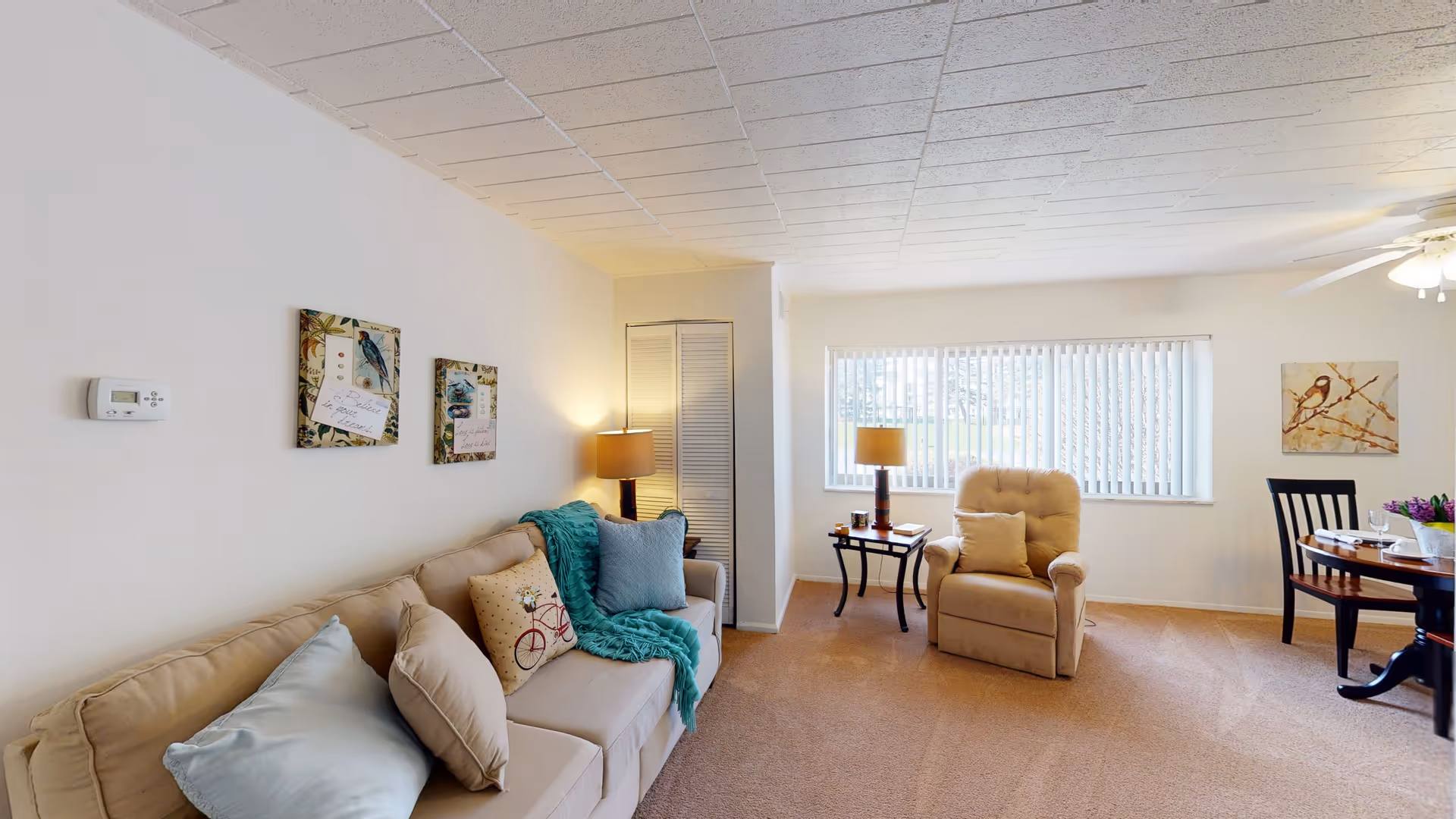 A cozy living room with a beige sofa adorned with multiple pillows and a teal throw blanket. There are two framed artworks on the wall above the sofa. A beige armchair with a matching pillow is positioned near a window with vertical blinds. Two side tables with lamps are placed beside the sofa and armchair. A round dining table with chairs is partially visible on the right side, set with plates, glasses, and a flower arrangement. The room has beige carpeting and a white ceiling with a ceiling fan.