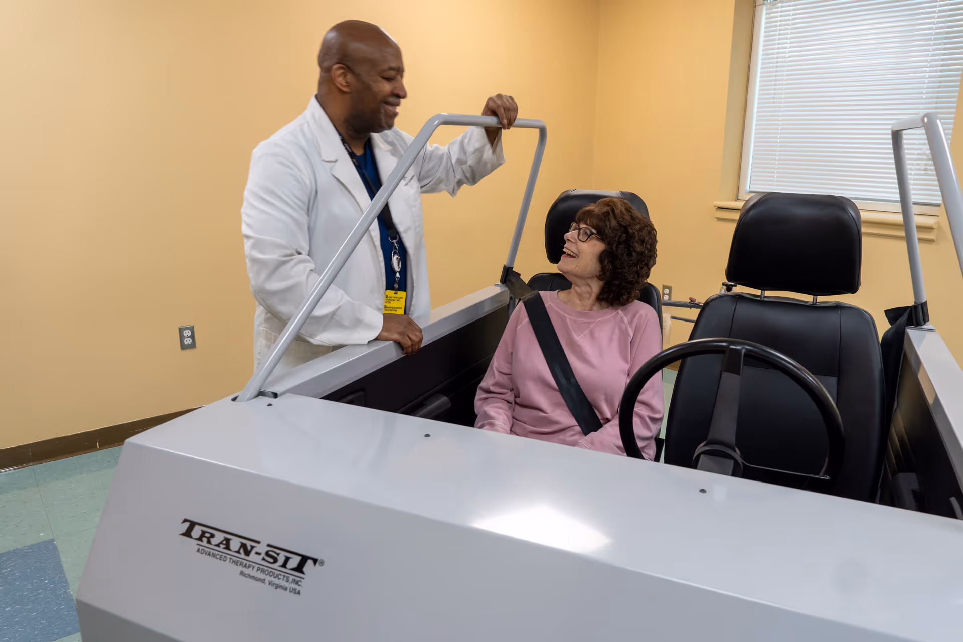 A healthcare professional in a white coat is standing next to an elderly woman seated in a driving simulation vehicle inside a room with beige walls and a window with blinds. The woman is wearing a pink sweater and glasses, and both are smiling and interacting.