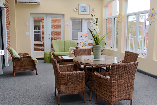 A bright indoor seating area with wicker furniture including a round glass-top table surrounded by four wicker chairs, a wicker loveseat with green cushions, and a wicker armchair. There is a green ceramic stool, a potted plant with white flowers on the table, and colorful artwork displayed near the window. Large windows and a glass door let in natural light, showing a brick exterior outside.