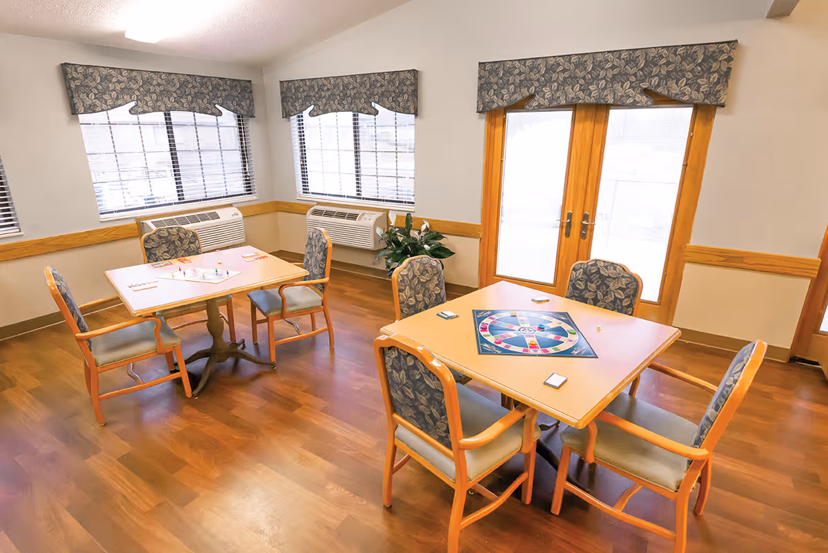 Well-lit interior activity room with two square tables and upholstered chairs set up for a board game near windows and glass doors.