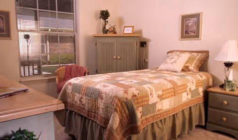A cozy bedroom with a single bed covered in a patchwork quilt. There is a wooden nightstand with a lamp and a small plant, a wooden cabinet against the wall, and a window with blinds letting in natural light. The walls have framed artwork and the room has a warm, inviting atmosphere.