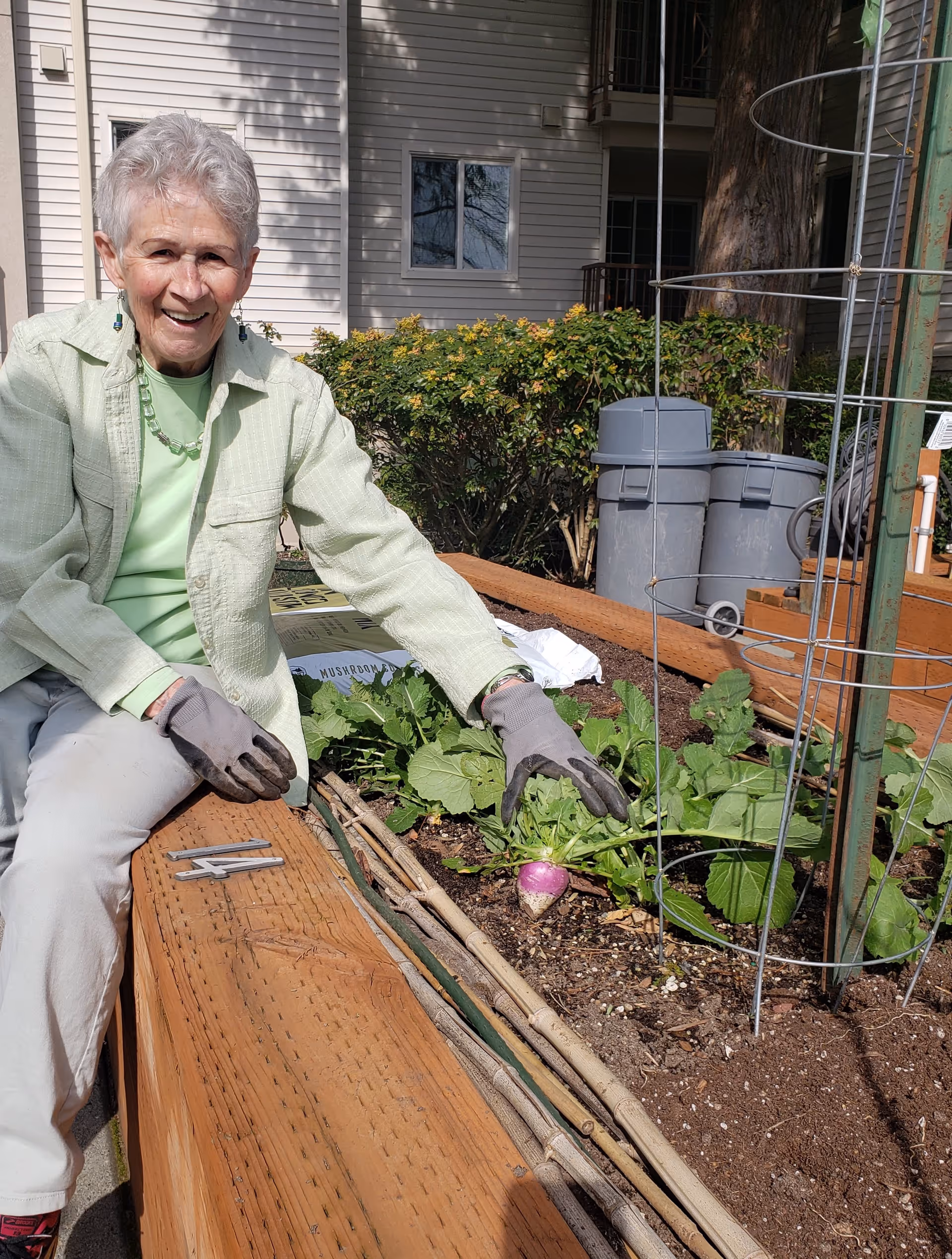 An elderly woman wearing gardening gloves and a light green jacket is sitting on the edge of a raised garden bed outdoors. She is smiling and holding a purple and white root vegetable, possibly a turnip, growing in the soil. Behind her are bushes, a building with windows, and two gray trash bins.